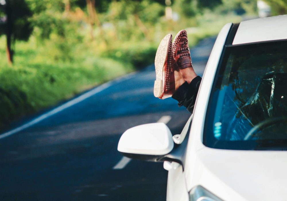 Person relaxing with their feet out of a car window while driving on a rural road surrounded by greenery.